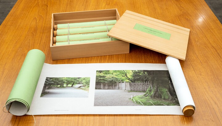A wood box containing green four green scrolls sits on a wood table; a fifth scroll in the foreground lies partially unrolled showing two photos of streets lines with trees and plants