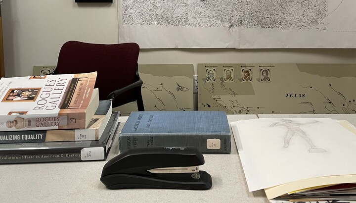 An office desk with (L-R) three library books stacked on top of each other; a cloth covered blue library book; a manilla file folder with papers sticking out of the end; in the foreground is a black Swingline stapler