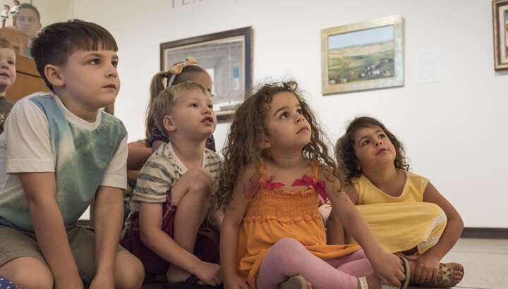 About six light-skinned children sit on the floor in a gallery; the four in the front are looking up at something out of the frame