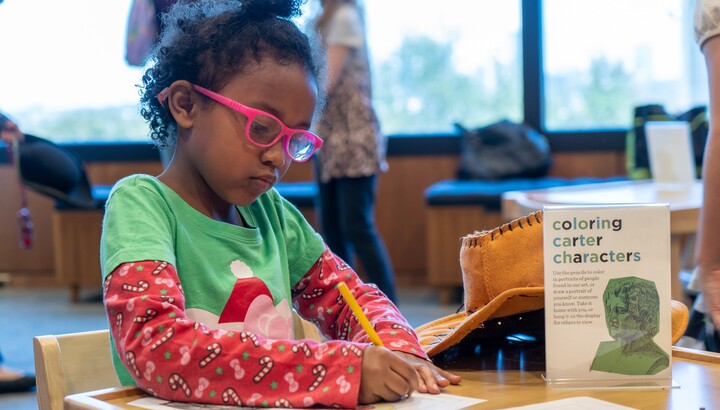 A dark-skinned child has her dark, curly hair pulled up on her head, wears pink glasses and a green and red t-shirt; the child sits at a desk with a pencil in her right had drawing on paper