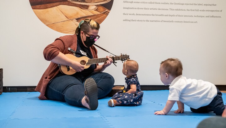 A woman sits on the floor of a gallery - right leg straight out and left leg folded in - playing a guitar; in front of her are two babies - one sitting and looking up at her, the other crawling toward her