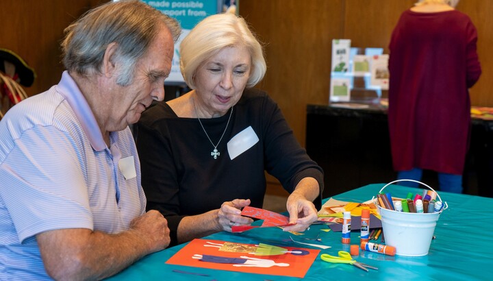 Two light-skinned elderly people sit at at table covered in art-making supplies; the man on the left looks closely at what the woman on the right is doing