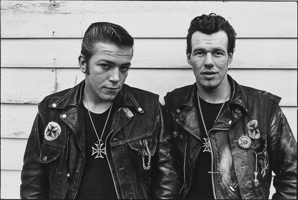 Two motorcyclists with leather jackets against building siding