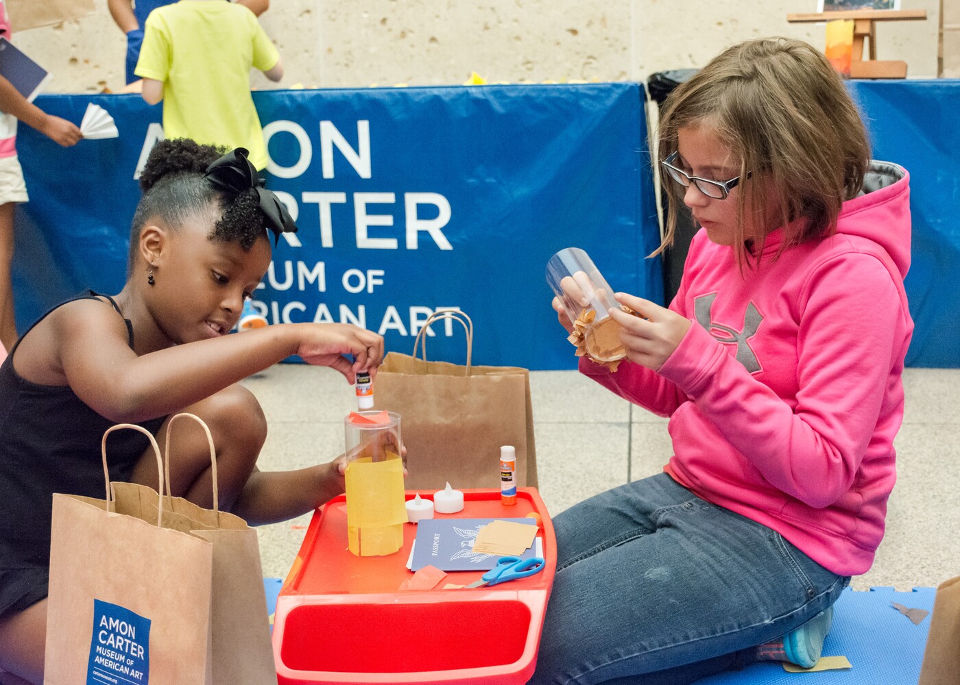 Two children are kneeling on either side of a small plastic red workbench during an art-making activity.