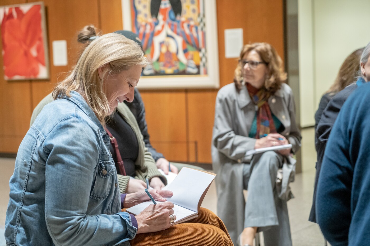 Three teachers are seated in a gallery with paintings hanging in the background. Each teacher has a journal in her lap, and the teacher in the foreground is smiling.