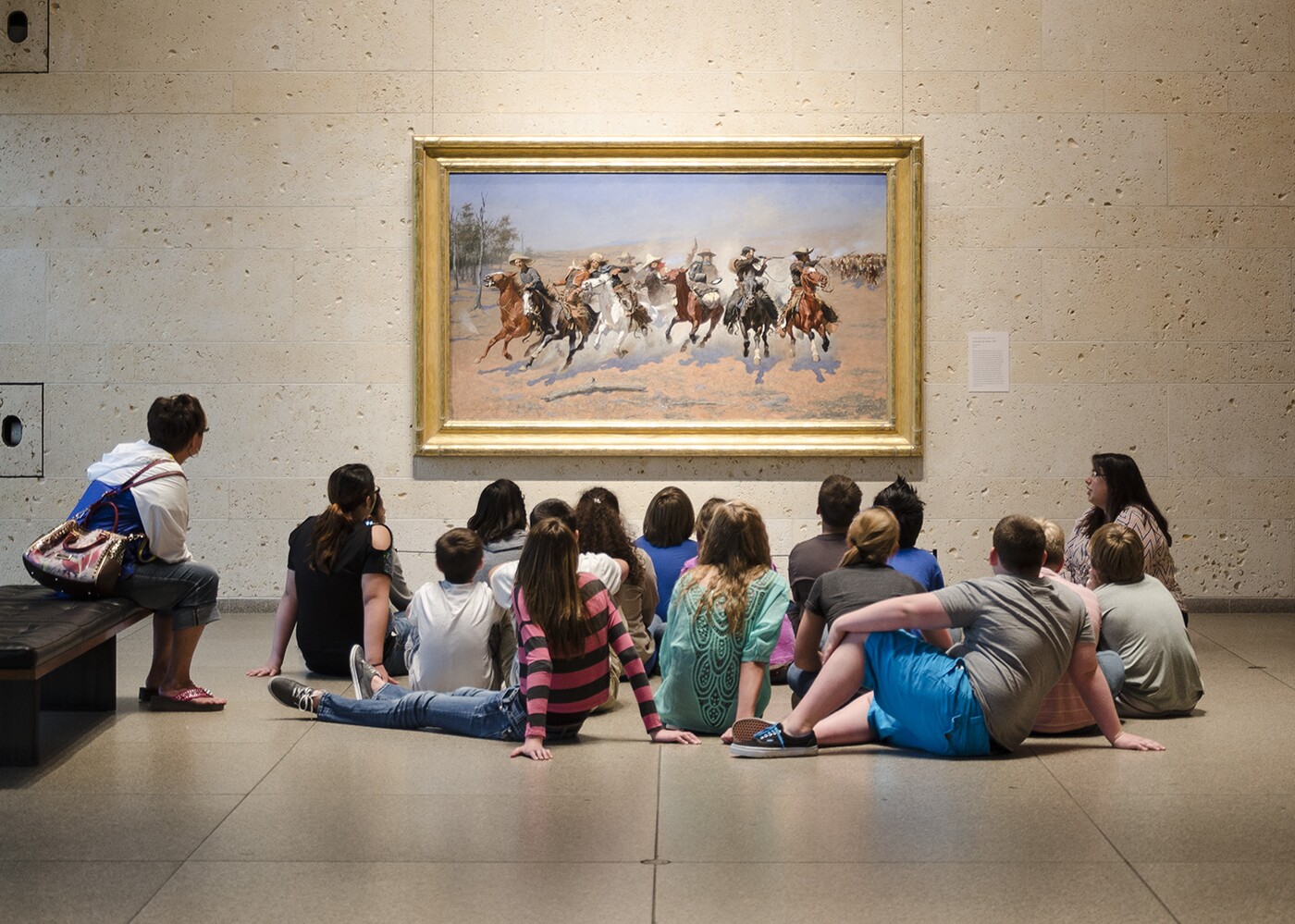 A group of middle-school students sit on the floor in front of a painting of horses and their riders galloping across the plains.
