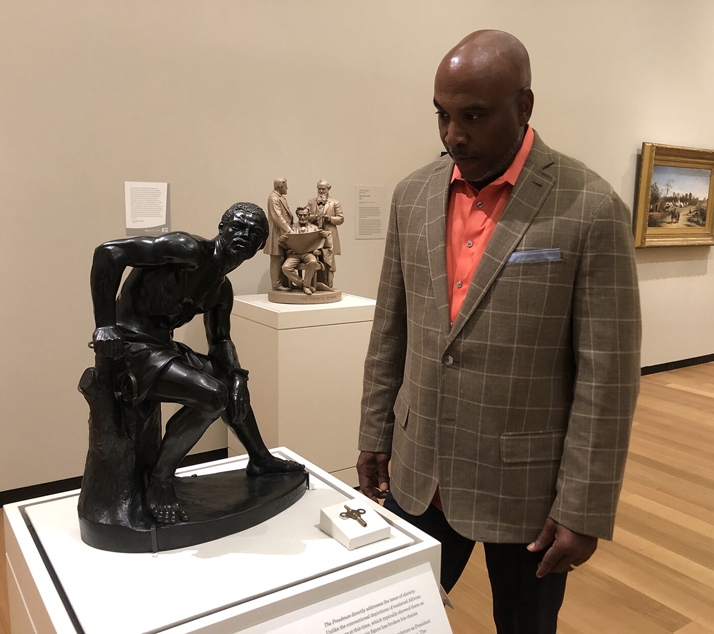 A dark-skinned man in a tan jacket looks at a bronze sculpture of a man sitting on a stump with his left arm resting on his left knee and his right hand, with shackles dangling from his wrist, on his right hip