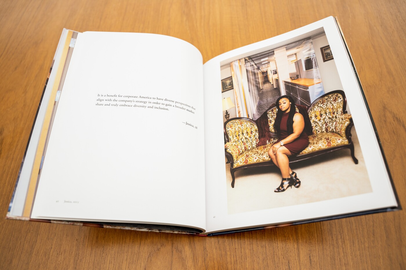 An open book lays on a wood table; the left page is white with three lines of text in the center (too small to read); the right page contains a full page photo of a medium-brown skinned woman sitting elegantly on a Victorian style couch