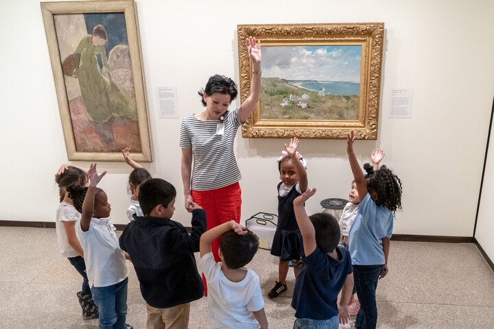A group of nine children stand facing an adult, who is standing in front of two hung paintings. All are raising hands.