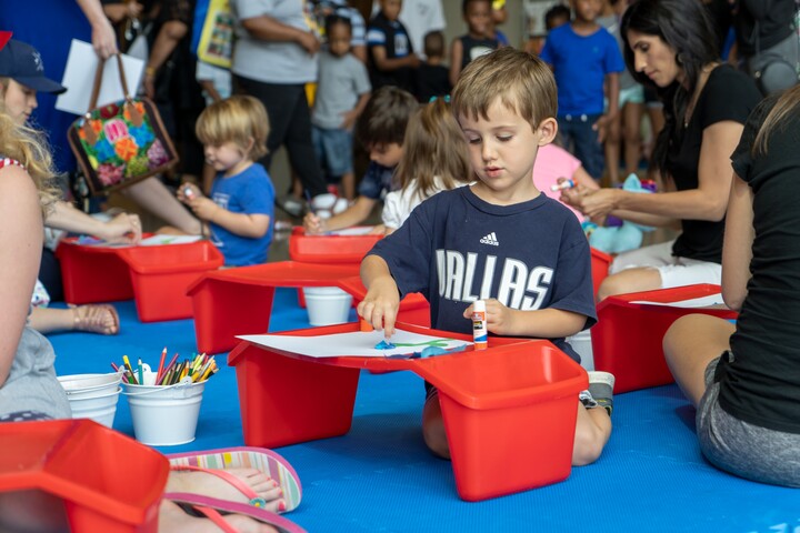 Many children are seated on the floor in front of red plastic workbenches during an art-making activity. The child in the foreground is dribbling paint onto a white piece of paper.