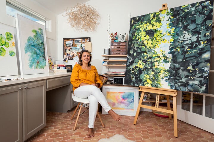 A woman wearing a gold shirt and white pants sits in a studio surrounded by paintings on the wall and on easels