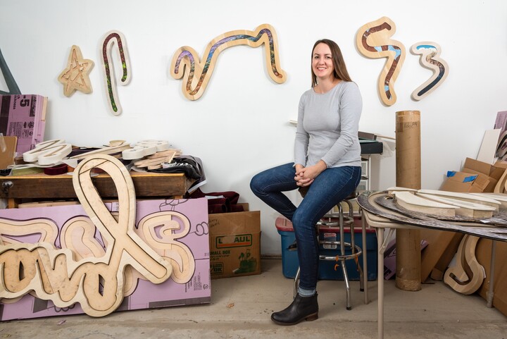 A woman in a gray top and jeans sits on a stool in an art studio with sculpture hanging on the walls and unfinished work on the floor