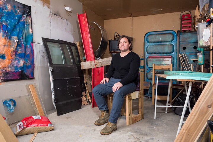 A man in a black shirt and blue jeans sits in a room full of sculptural and found objects