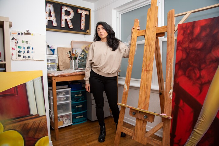 A woman in a tan shirt and black pants stands next to an empty easel in an art studio
