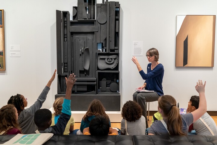 Approximately 10 students sit on a gallery floor, some with their hands up, as a Gallery Teacher talks about a large black sculpture in front of them