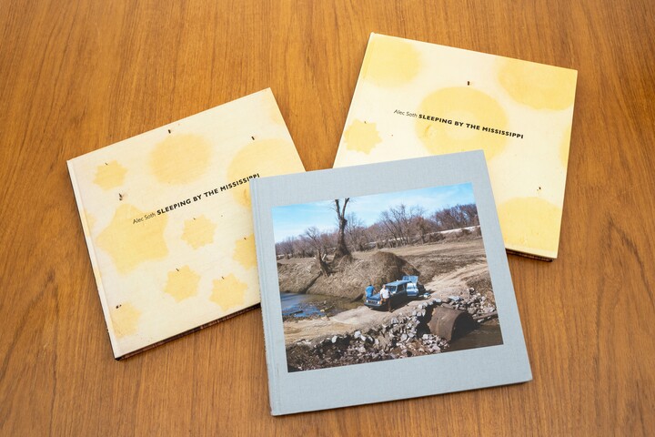 Three books lay on a wood table; two of them them have light yellow covers with dark yellow stars and circles; the one in the foreground features a photo of two men leaning on a car that is parked on a culvert