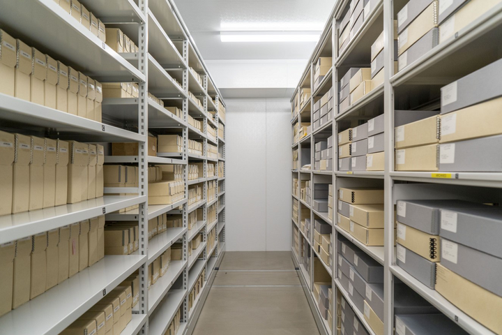 A view down a row of metal shelves on which gray and cream-colored archival boxes are stacked neatly