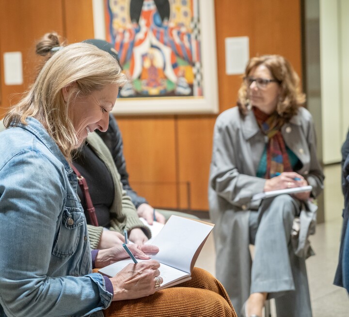 Three teachers are seated in a gallery with paintings hanging in the background. Each teacher has a journal in her lap, and the teacher in the foreground is smiling.