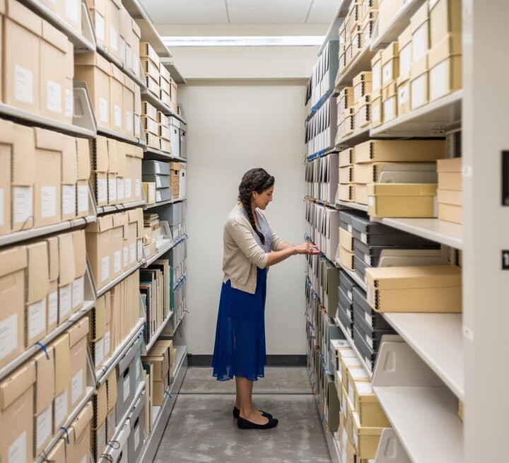 Rows of archival shelves are on either side, with a woman in the middle reviewing material.