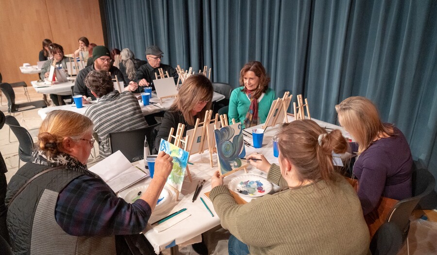 Several teachers are seated around four rectangular tables. Each teacher has a small wooden easel in front of them, on which is mounted a small canvas for painting.