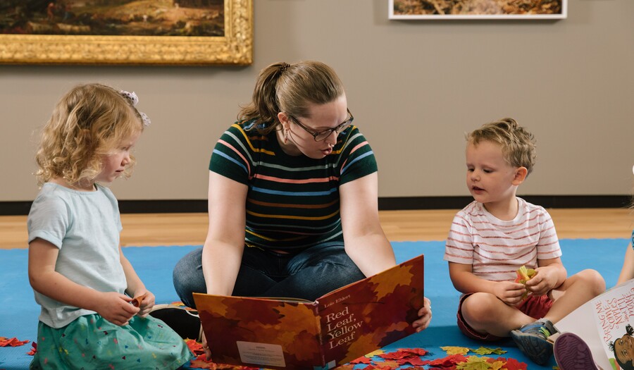 teacher reading a book to a little girl and little boy in front of a landscape painting
