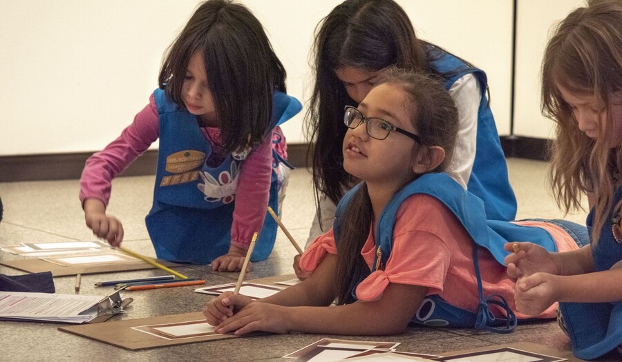 Four girls in Girl Scout daisy uniforms are on the floor of a gallery, three look at a sketching project and one looks up