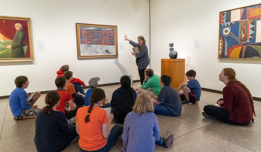 Woman standing in a gallery motions toward painting while group of children and teens sits on the floor
