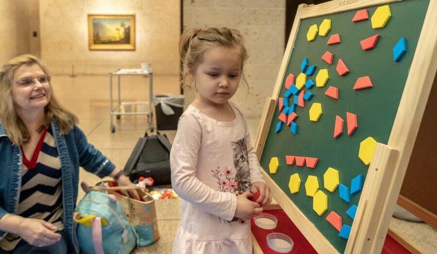A small child stands at an easel with colored shapes arranged on it as a woman seated on the floor looks on