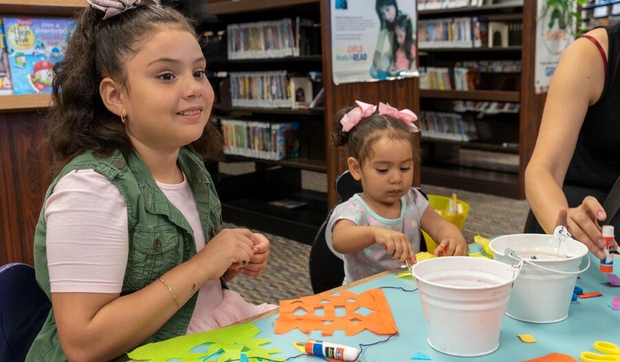 Two children sit at a table strewn with art supplies; they are each working on their own projects