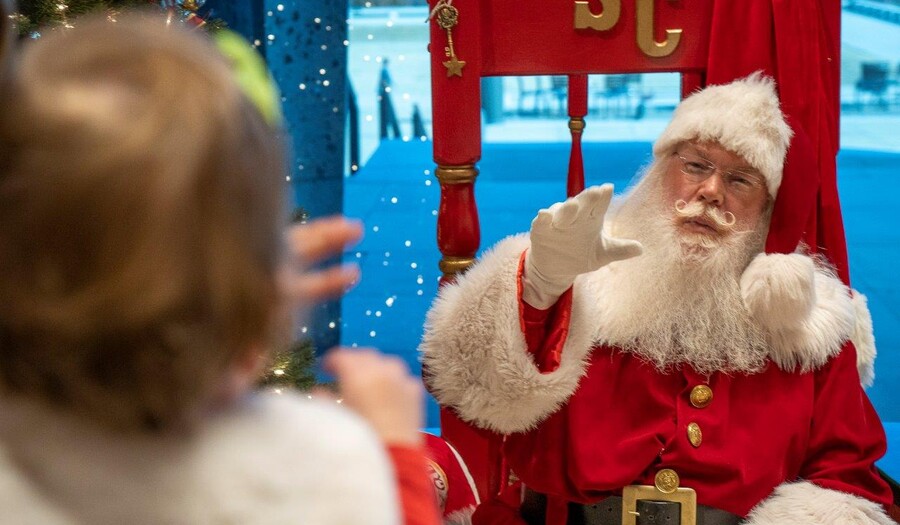 Santa Claus waves to a small child in the foreground who is being held by someone out of view