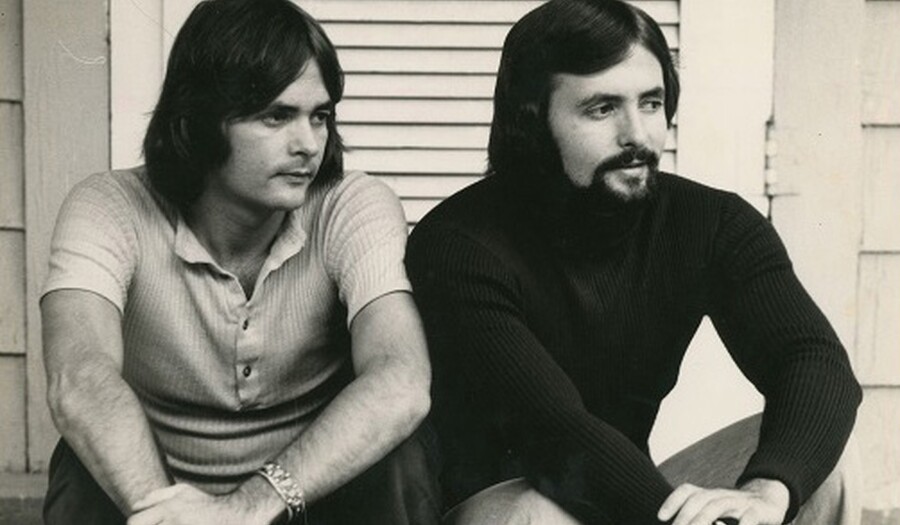 Black and white photo of two light-skinned men with dark hair sitting on what looks like a porch; the man on the left is wearing a light-colored, short-sleeved polo and has his arms resting on his knees with his left hand resting on his right wrist; the man on the right is wearing a black turtleneck, he has a beard and mustache and is sitting in a similar position; they both look off to their left