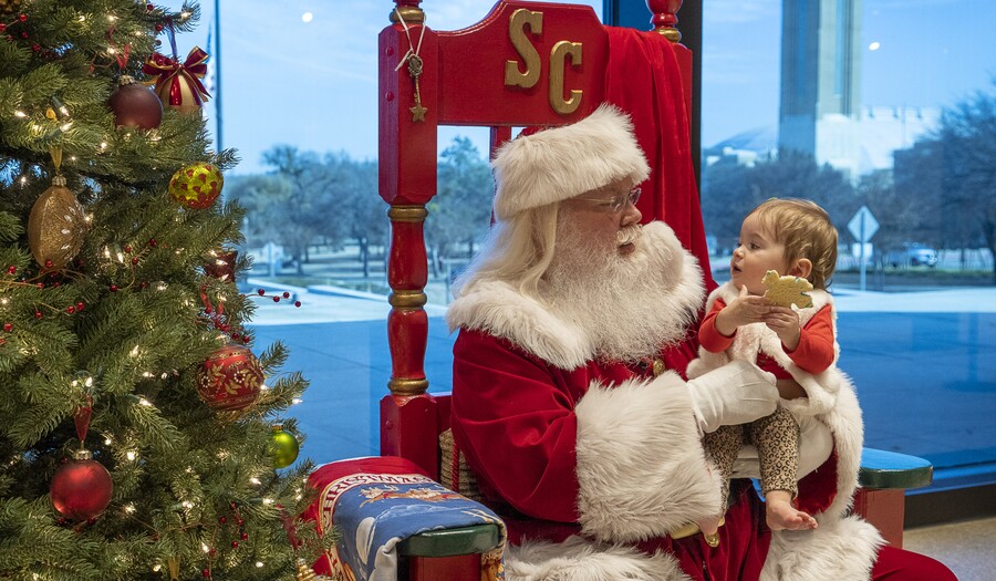 Santa Claus sits in a red throne-like chair in front of a window and to the left of a Christmas tree; In his left arm he holds a toddler who looks at him and holds a cookie in her hands