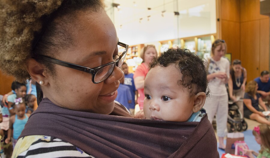 A dark-skinned woman with glasses looks lovingly at a dark-skinned baby in a baby sling on her chest