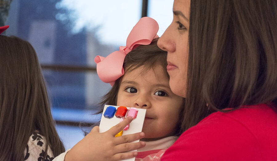 A close-up of a toddler with a large pink bow in her brown hair sitting in a woman&#039;s lap; the child holds a a white card with blue, red, and pink color swatches on it in her right hand covering part of her mouth
