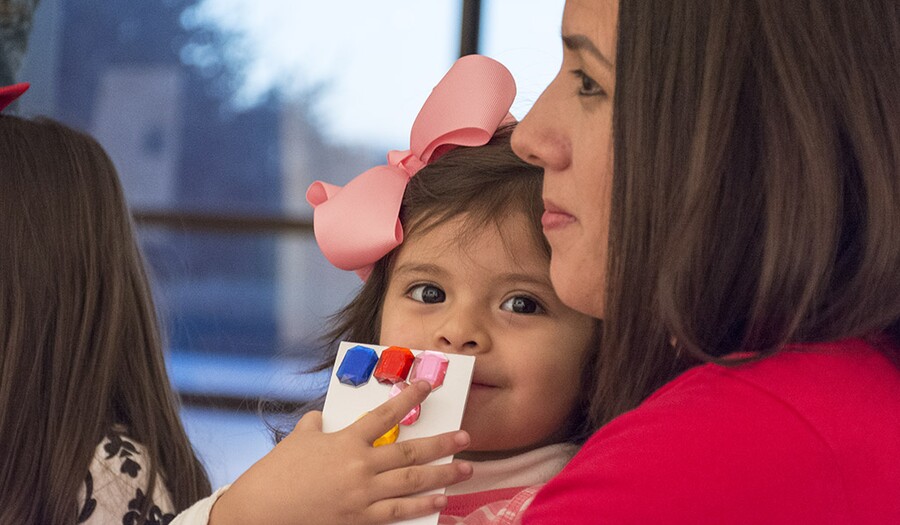 A close-up of a toddler with a large pink bow in her brown hair sitting in a woman's lap; the child holds a a white card with blue, red, and pink color swatches on it in her right hand covering part of her mouth