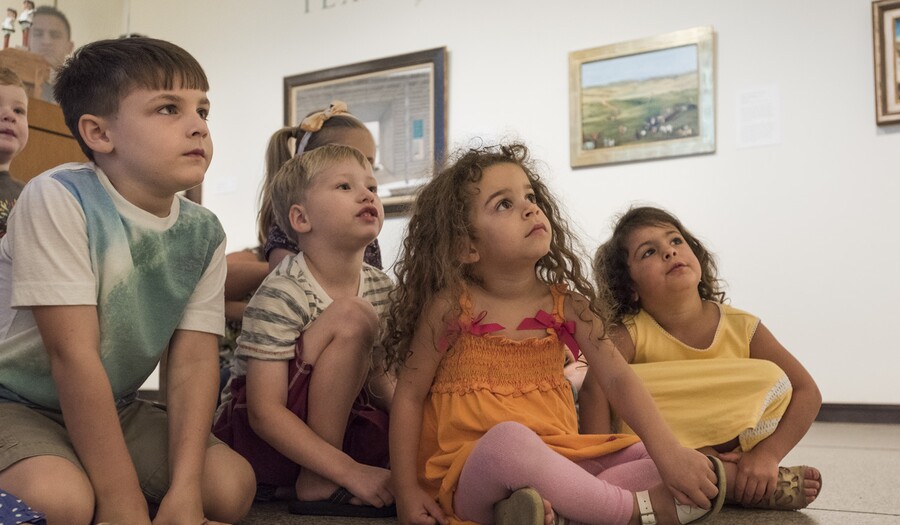 About six light-skinned children sit on the floor in a gallery; the four in the front are looking up at something out of the frame