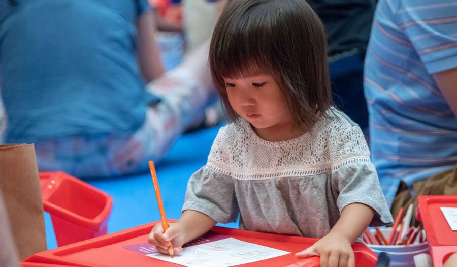 A small child with dark hair cut in a bob leans over a red desk on which she is drawing on a piece of paper