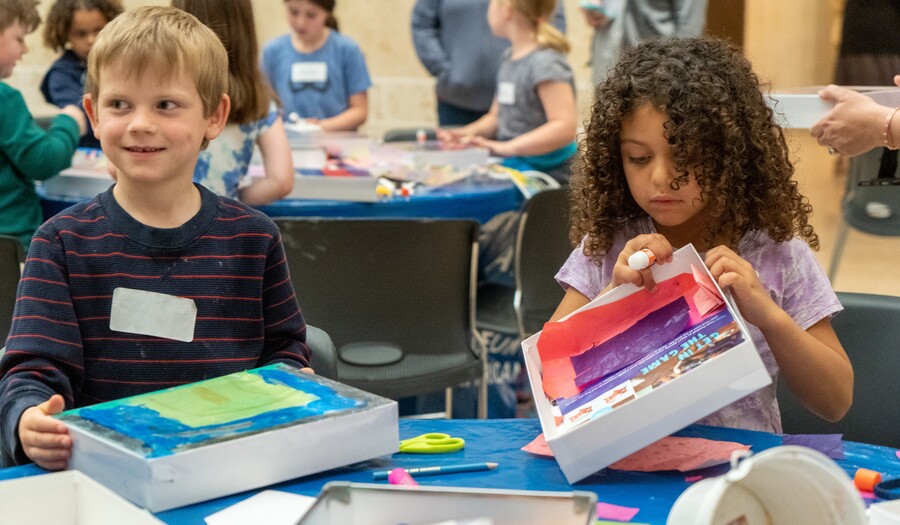 Two children sit at a table making art; the light-skinned blond child on the left holds a white box with blue and green paper on it and looks mischievously to his right; the medium-skinned child on the left has medium-lenght dark curly hair and is holding a white box with purple and pink paper on it
