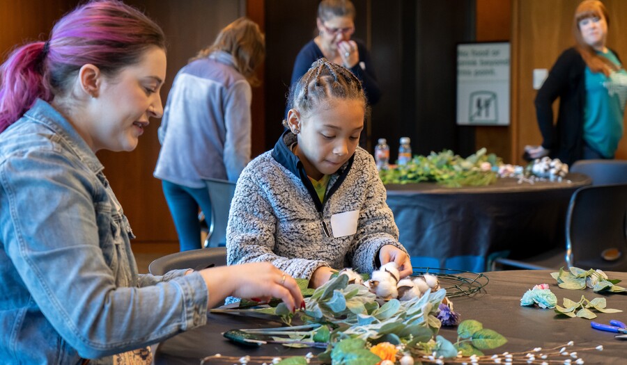 A light-skinned adult with purple hair pulled into a ponytail sits at a table with a medium-skinned elementary-age child with braided hair; they are working on an art project together