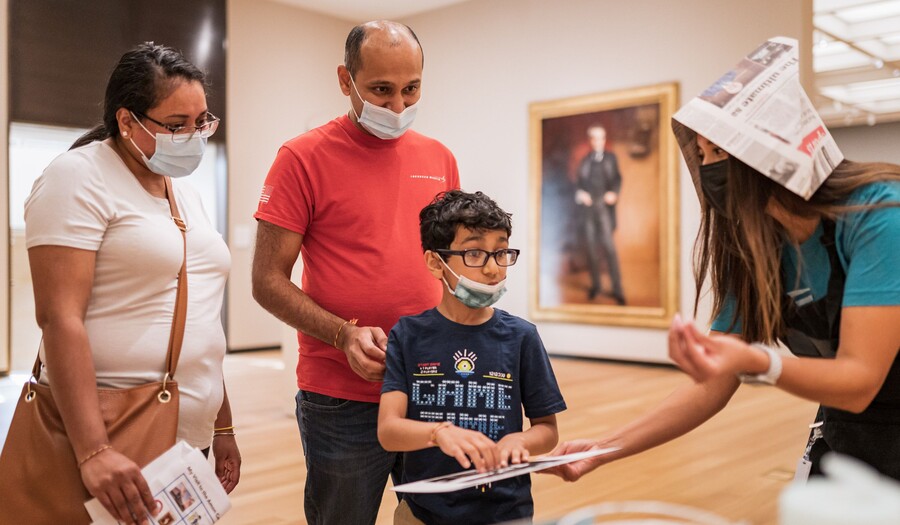 Three adults and a child in a gallery, all wearing masks; (L-R) a woman in a white shirt, tan pants, and long dark hair pulled back in a pony tail watches the child in front of her; a man in a salmon-colored shirt and jeans stands behind the child also watching; the child has short dark hair, wears glasses, and a t-shirt reached his hands out to touch a tablet that is held by a woman on the far left wearing a newspaper hat
