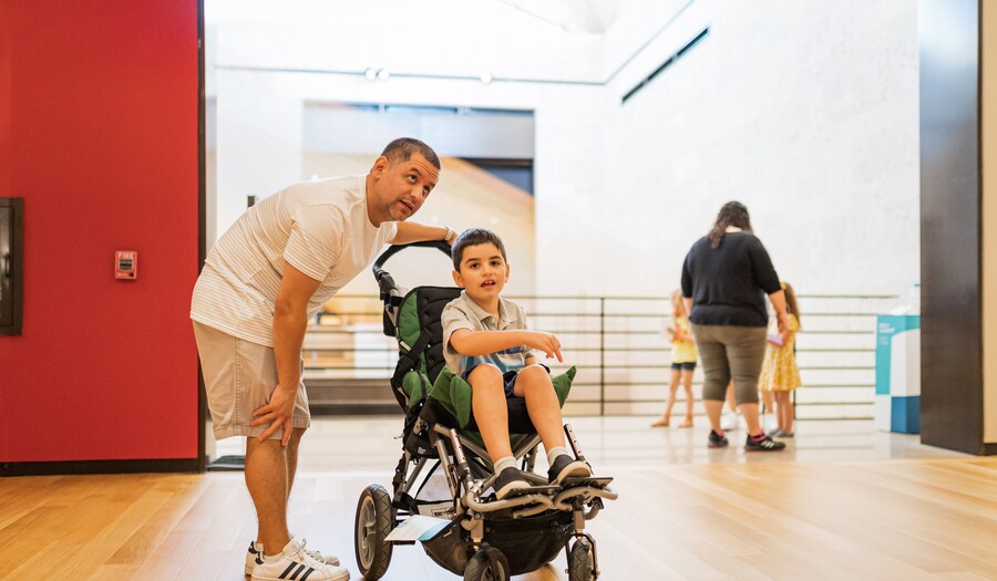 A light-skinned adult with close-cropped dark hair and a short goatee leans over an older child with short dark hair in a green stroller; they are in a museum gallery and the adult is looking up at something