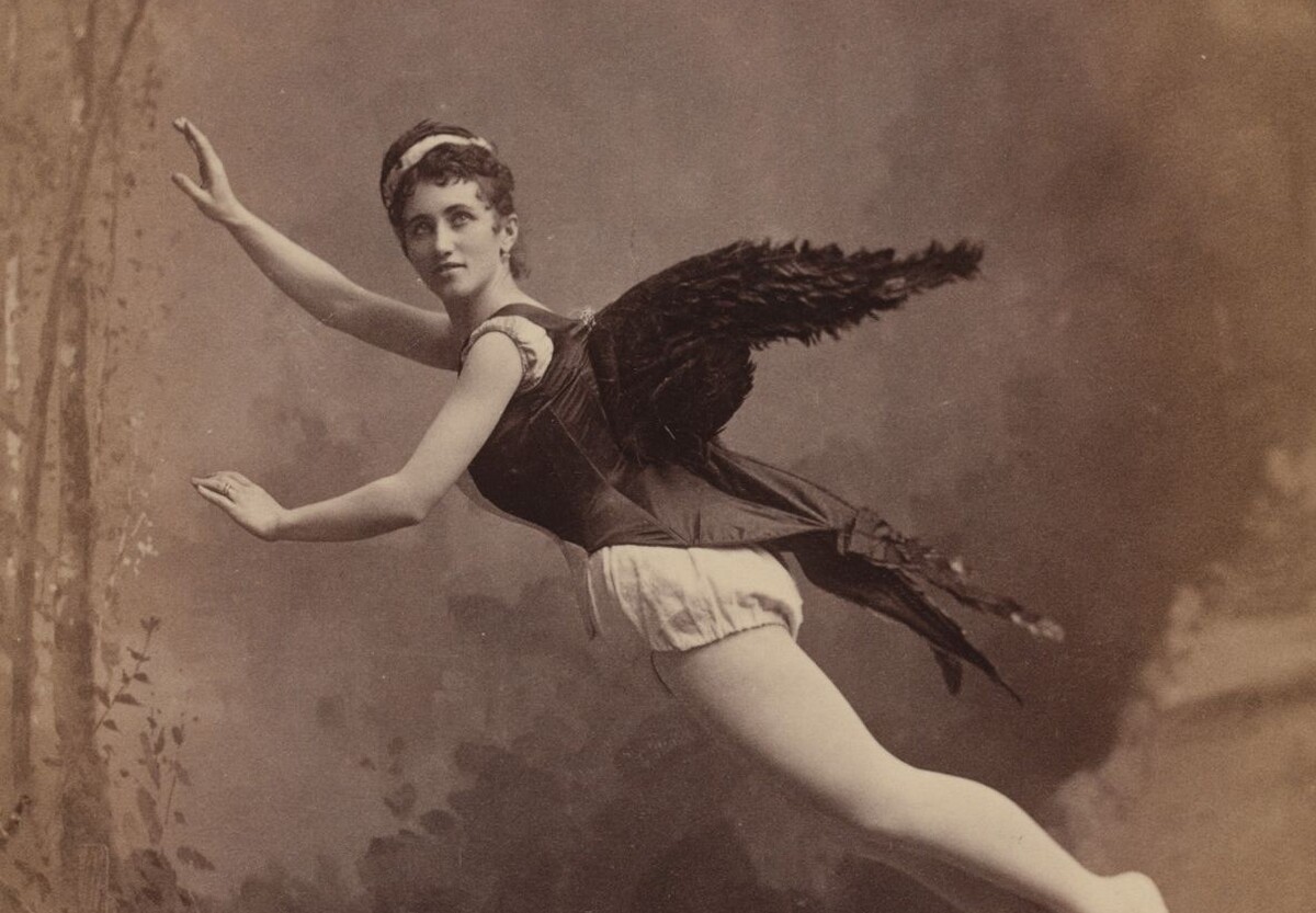 Black-and-white photograph from the 1880s of a woman wearing a dark corset with black feathered wings attached to the back and posed as if she is flying